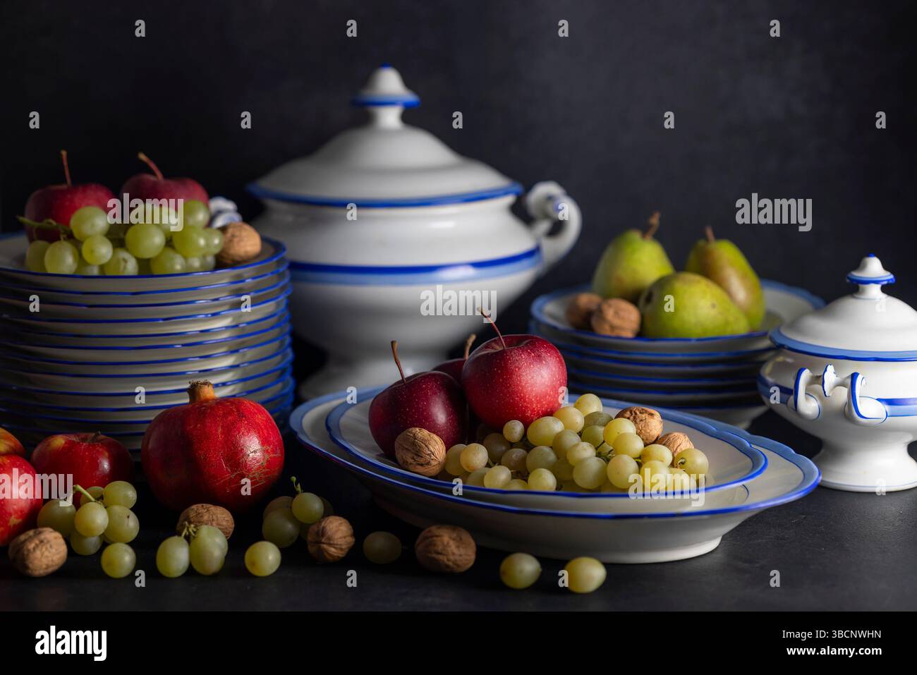 Antiche stoviglie in porcellana blu e bianca con mele, uva, melograno, pere e noci su sfondo scuro che compongono una natura morta Foto Stock