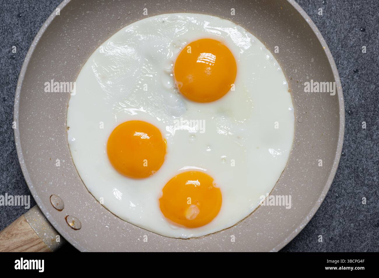 , Deutschland, 21.05.2025, Frische Spiegeleier in heller Bratpfanne mit Holzgriff, auf dunklem Untergrund. Studioaufnahme, Draufsicht. Symbolbild für Foto Stock