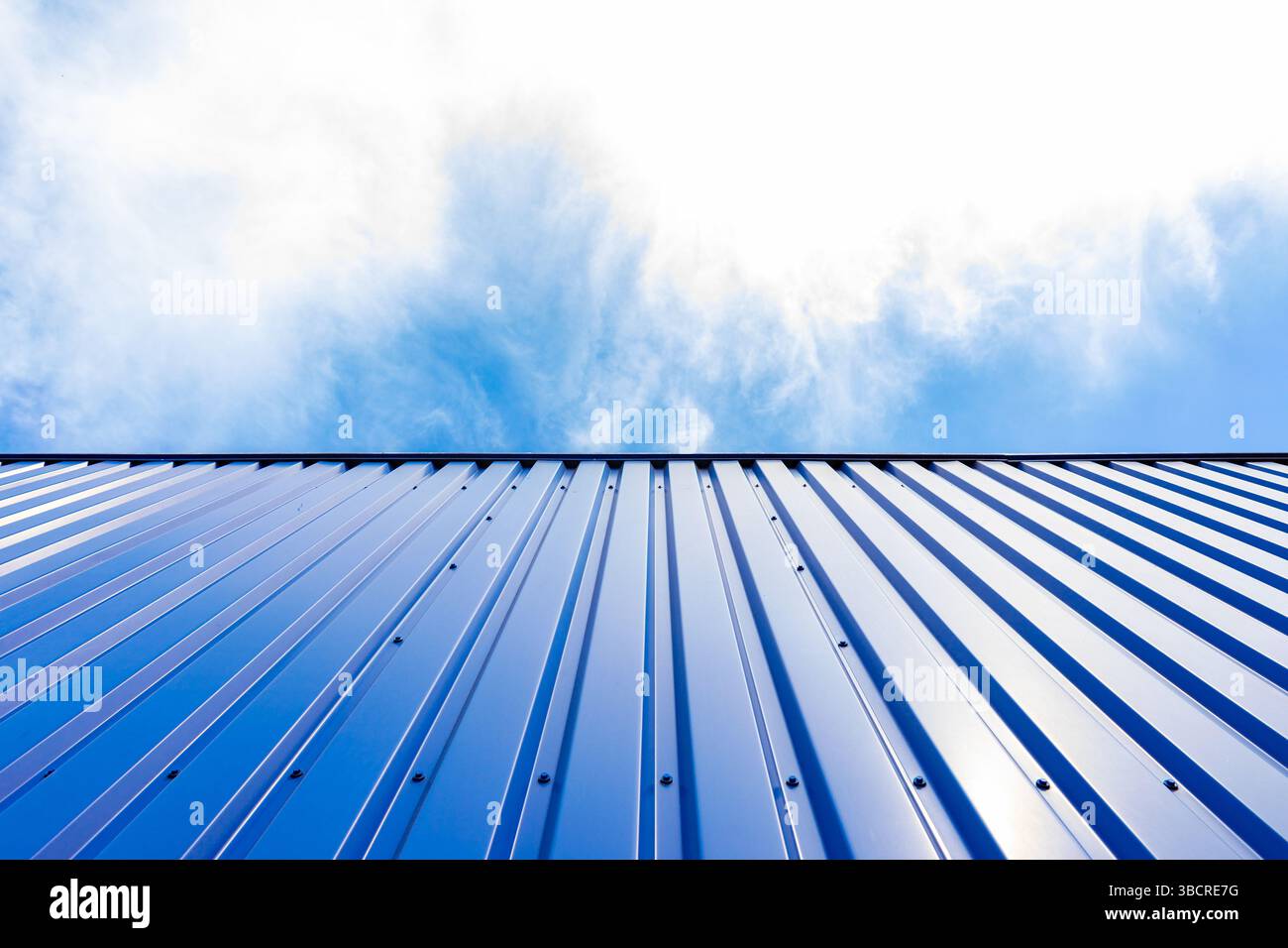Vista ad angolo basso di una parete in metallo ondulato blu contro un cielo luminoso con nuvole morbide, ideale per temi industriali o architettonici. Foto Stock