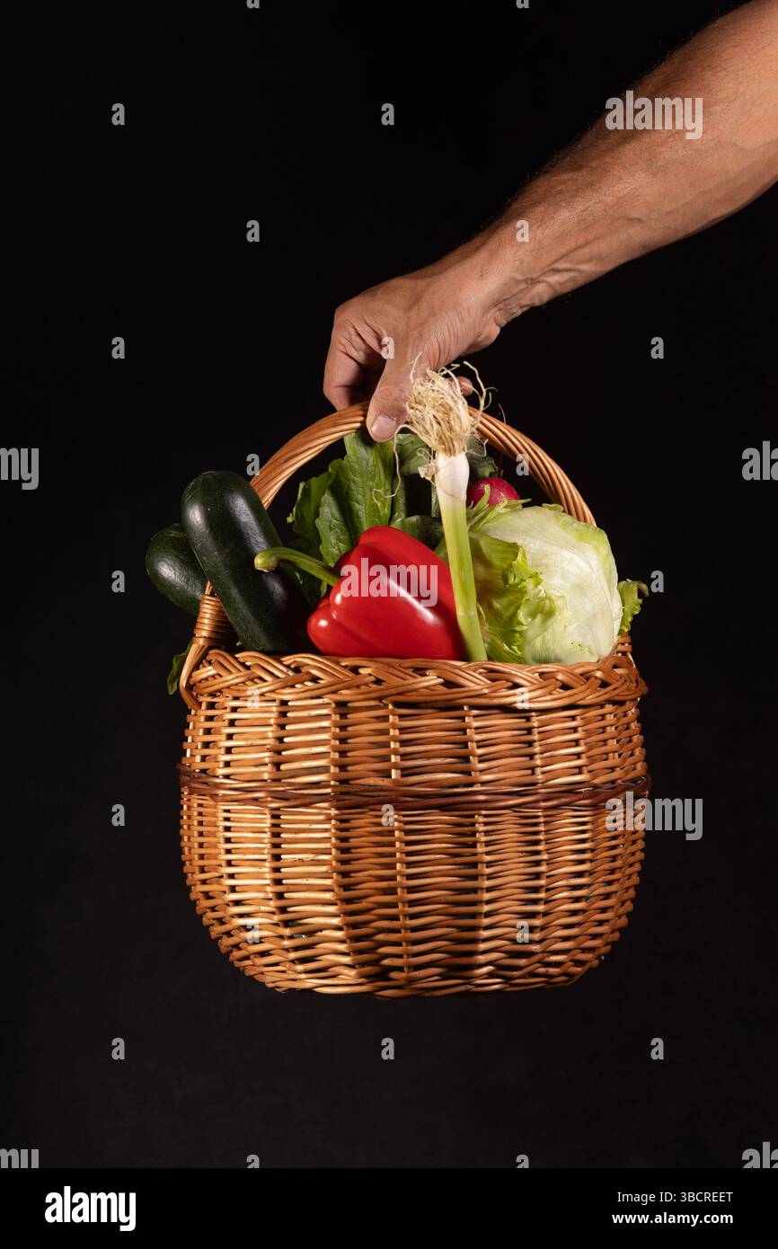 Una mano regge un cesto di vimini riempito con varie verdure su sfondo nero. Il cestino contiene prodotti vivaci, tra cui peperoni rossi Foto Stock