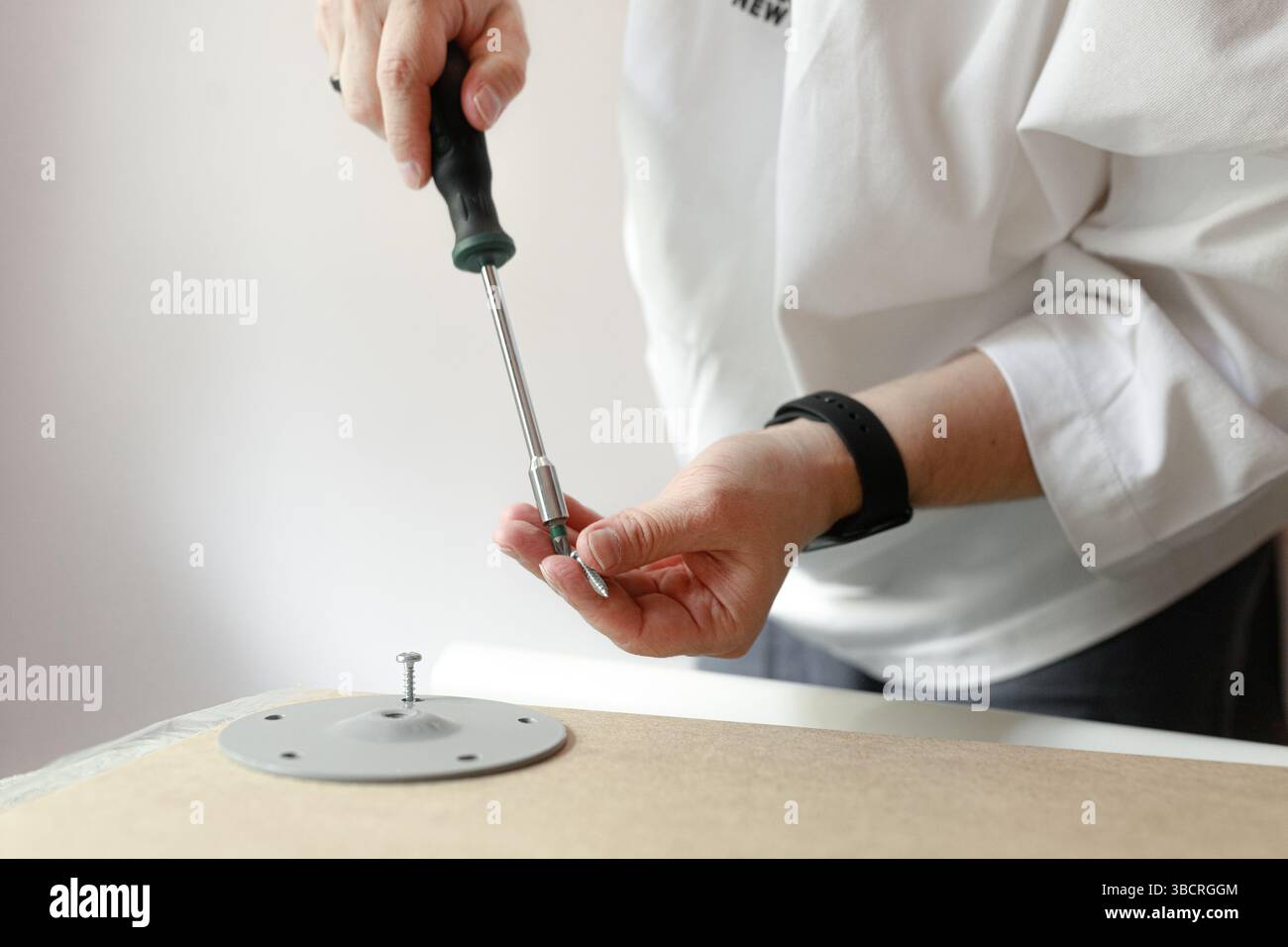 Persona che prepara una vite e una punta per fissare una piastra metallica rotonda su un pannello di legno utilizzando un cacciavite. Foto Stock