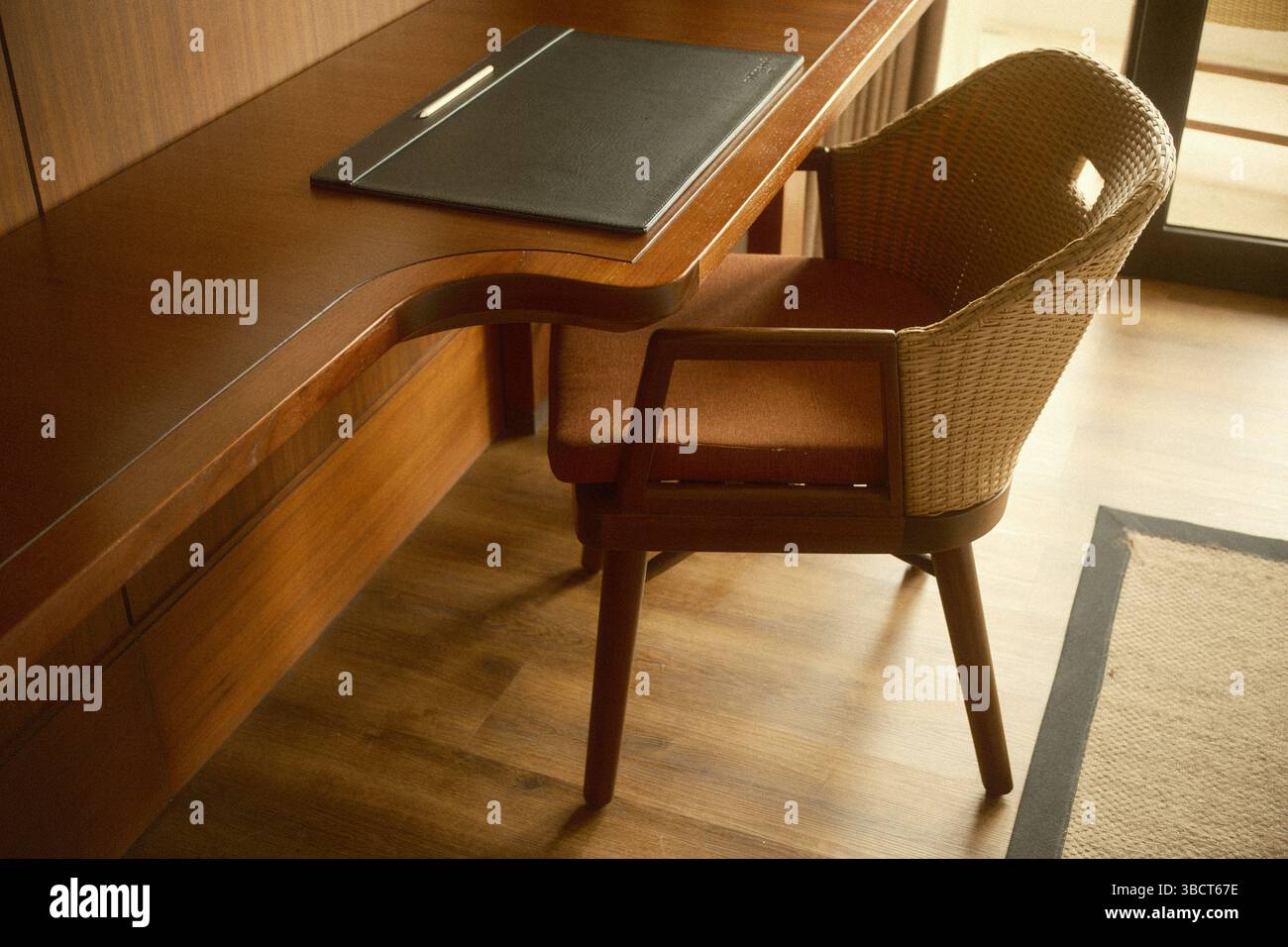 Uno spazio di lavoro con una scrivania in legno, una sedia in rattan e un tappeto da scrivania in pelle Foto Stock