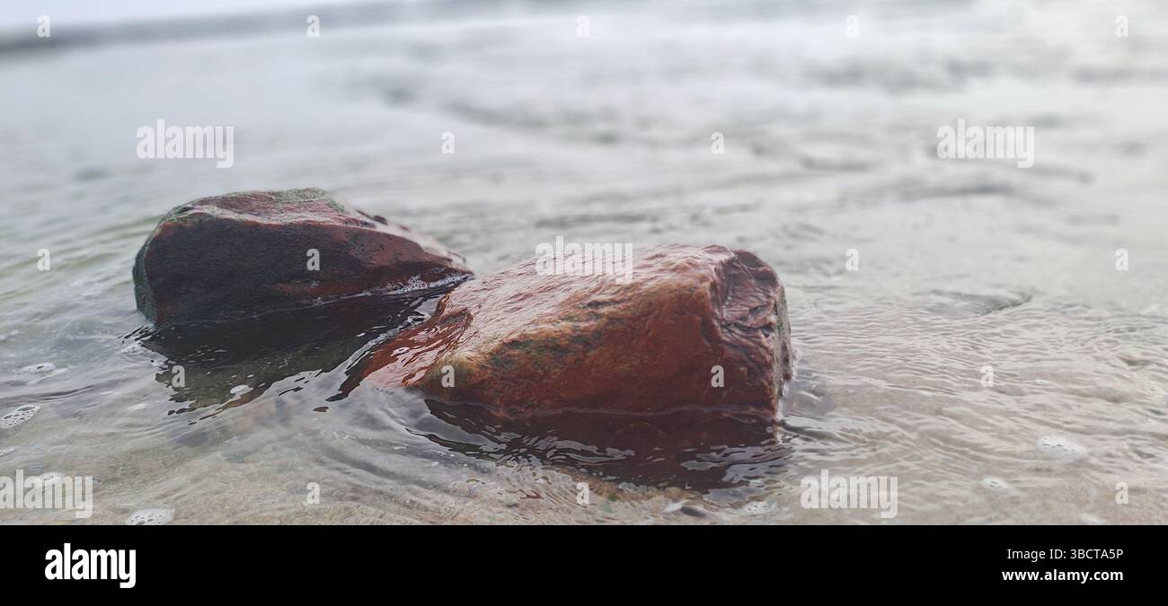 Onde dolci che scivolano su due rocce rossastre su una spiaggia sabbiosa Foto Stock