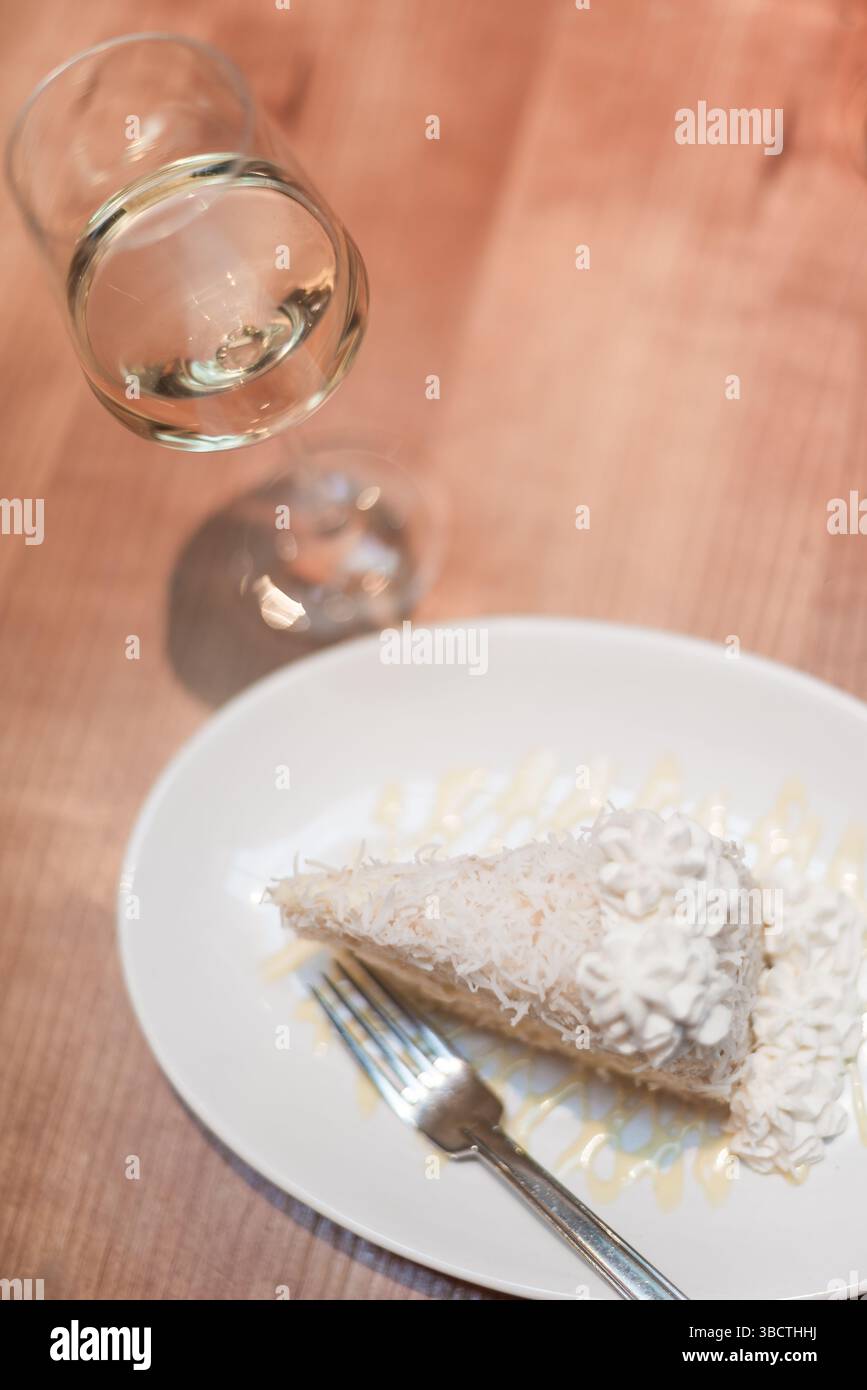 Torta al cocco e abbinamento di vino bianco // Una fetta generosa di torta al cocco si siede accanto a un bicchiere di vino bianco su un piano di legno semplice in un elegante ambiente da ristorante. La torta a strati, con la sua glassa bianca e i fiocchi di cocco, crea un contrasto sorprendente con la superficie in legno naturale. Il bicchiere di vino di cristallo contenente vino dorato pallido completa il dessert, dimostrando un abbinamento sofisticato che bilancia la dolcezza della torta con l'acidità del vino. Foto Stock
