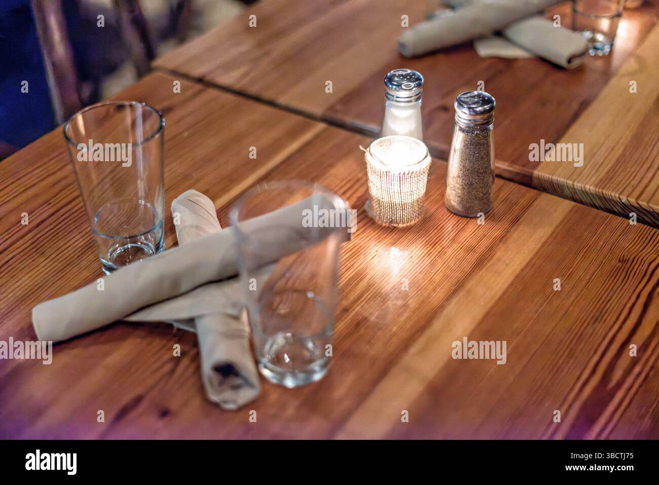 Tavolo della Taverna con bicchieri e candela // Un tavolo in stile taverna disposto in modo informale ma invitante su un tavolo in legno massiccio. L'ambiente include bicchieri vuoti in attesa di bevande, posate avvolte in tovaglioli per una facile manutenzione, saline e pepe per condire e una candela votiva che fornisce una calda illuminazione ambientale. La superficie rustica in legno aggiunge calore e carattere all'atmosfera informale. Foto Stock