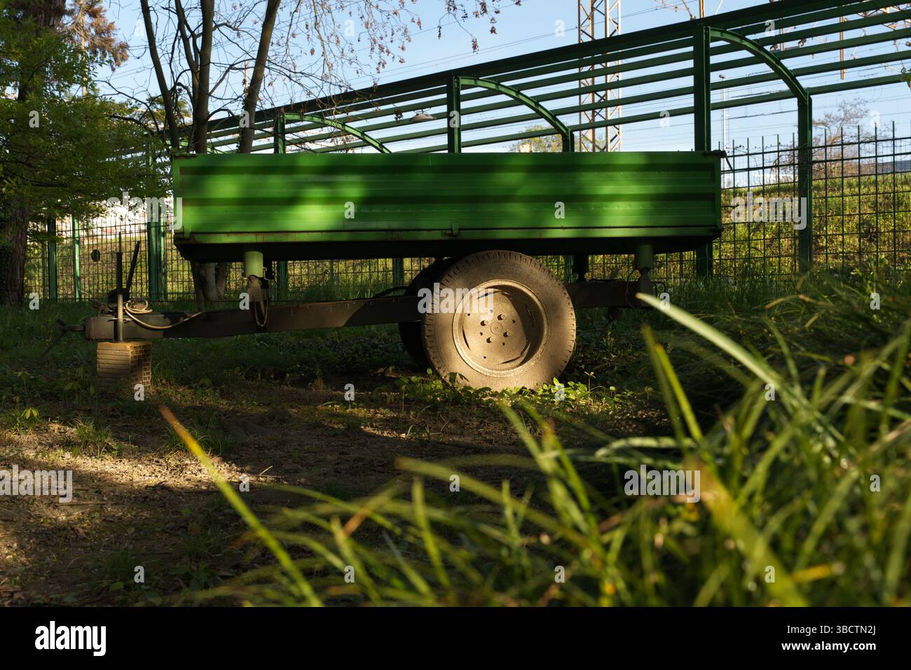 Un rimorchio verde con è parcheggiato sull'erba vicino a una recinzione metallica e agli alberi. La luce del sole proietta ombre, evidenziandone la robustezza Foto Stock