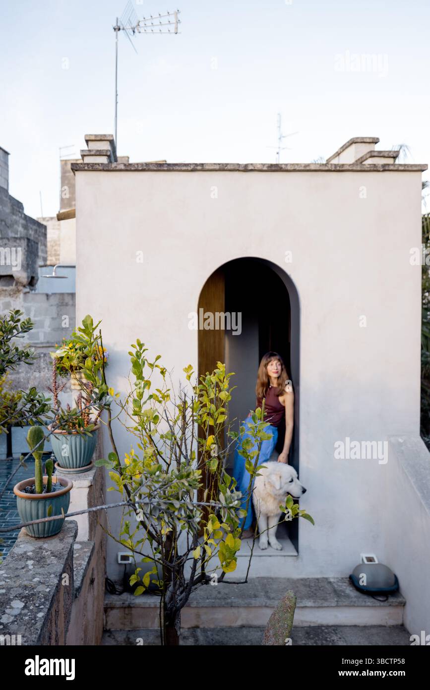 Una donna e il suo cane bianco escono su un giardino sul tetto circondato da piante in vaso e architettura in pietra, evocando un tranquillo stile di vita mediterraneo e un legame con la natura Foto Stock