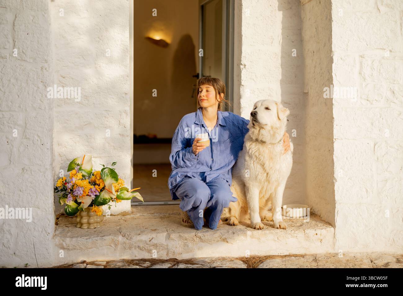 Un momento sereno condiviso tra una donna in lino blu e il suo leale cane bianco, godendosi un'alba tranquilla alle porte di una casa tradizionale di trullo in Puglia, Italia Foto Stock