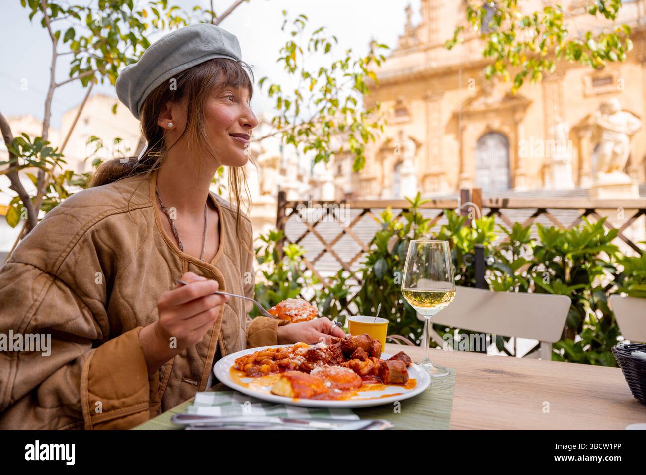 Una donna sorridente si diverte a gustare un abbondante pranzo siciliano con gnocchi, polpette e salsa di pomodoro, abbinato a vino bianco, in un ristorante all'aperto di fronte alla Cattedrale di San Pietro a Modica Foto Stock