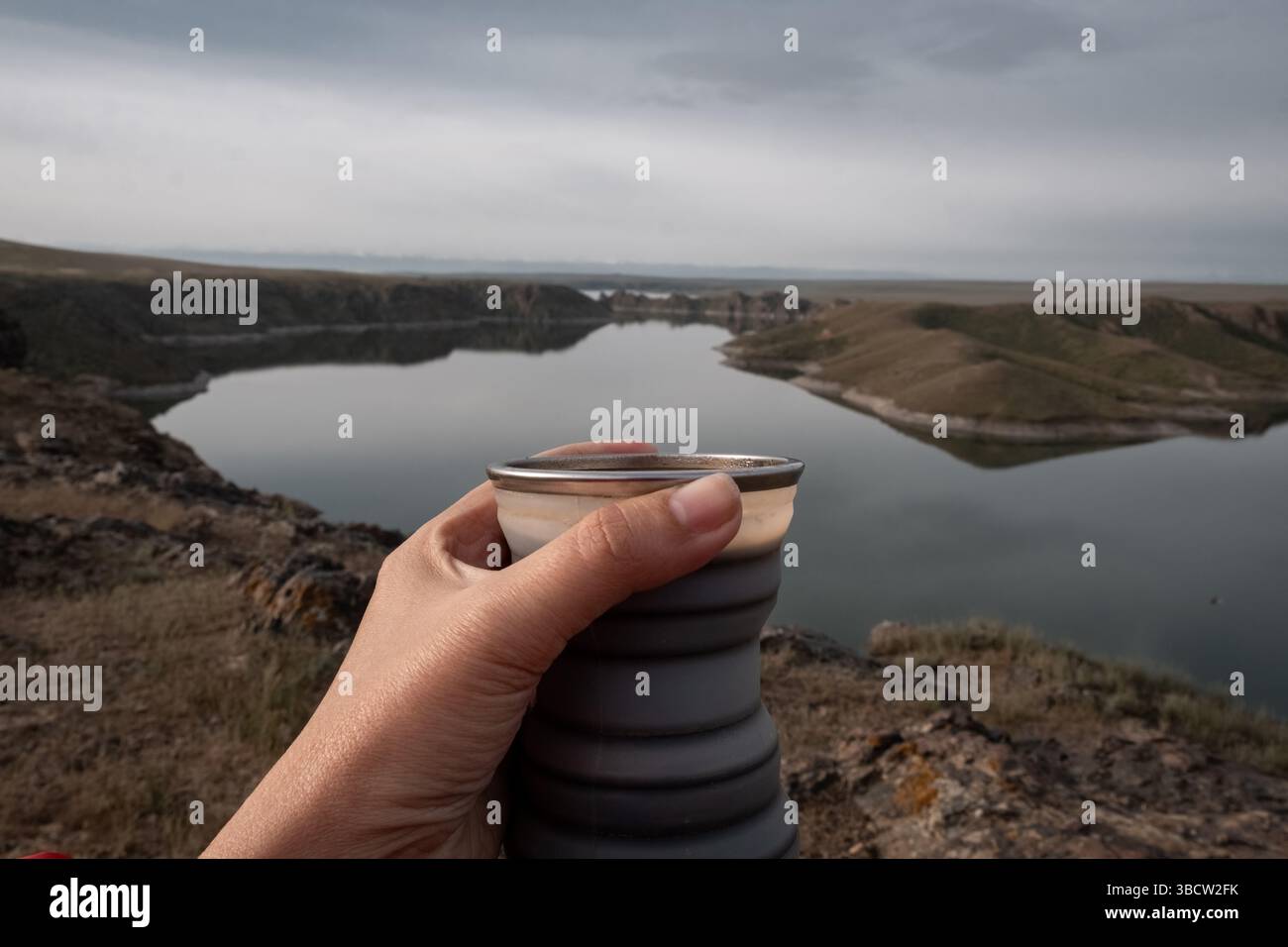 Primo piano di una donna che tiene una bevanda calda in una tazza pieghevole in silicone per turisti sullo sfondo di un lago autunnale con rocce. Kurty o Kurta Res Foto Stock