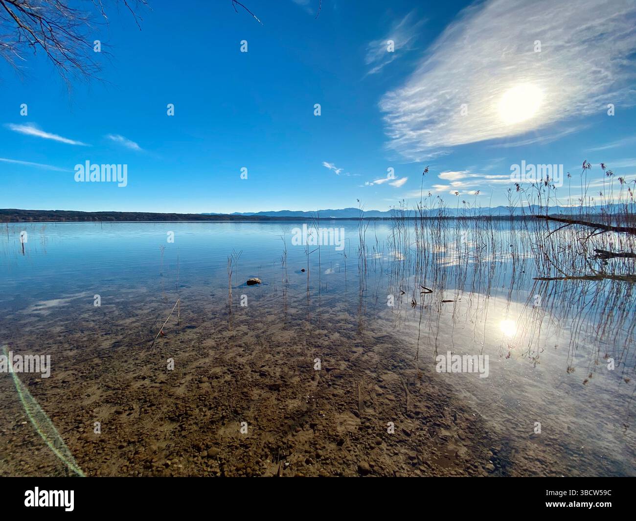 Lago blu sereno che riflette un sole luminoso e nuvole irregolari. L'acqua è limpida, rivelando un fondo sassoso e lunghe canne sottili che si estendono verso l'alto. Foto Stock