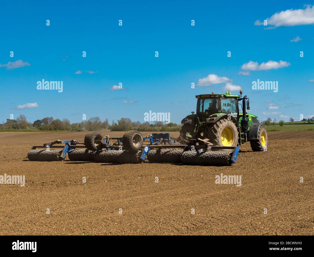 Trattore agricolo John Deere di grandi dimensioni verde che tira il rullo di coltivazione in un campo agricolo appena arato nel Leicestershire ad aprile, Inghilterra, Regno Unito Foto Stock