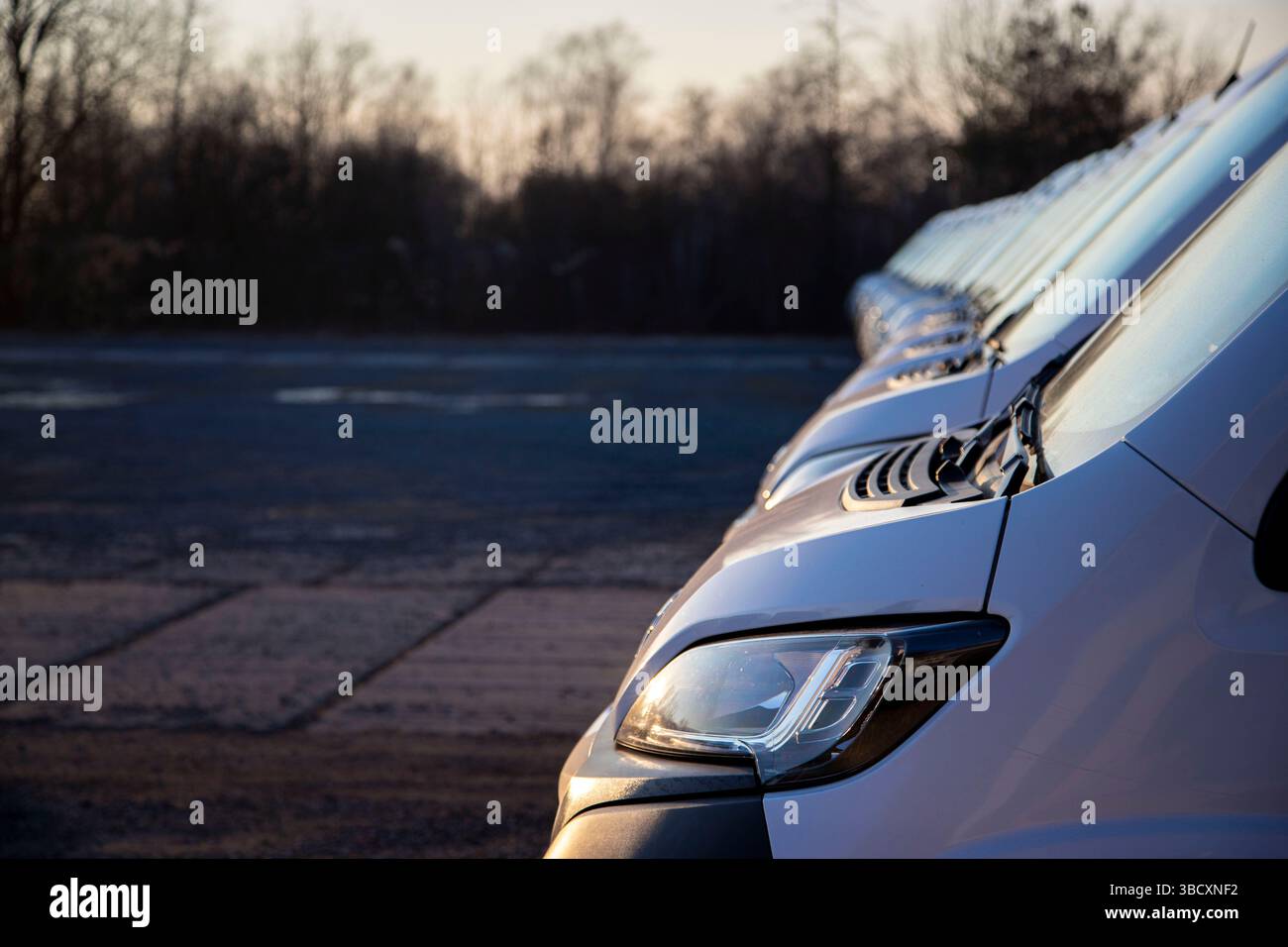 Lato e parte anteriore di un furgone con pannelli in una lunga fila di furgoni bianchi identici pronti per la consegna al cliente. Faro, cofano, parafango, paraurti. Una linea di furgoni. Foto Stock
