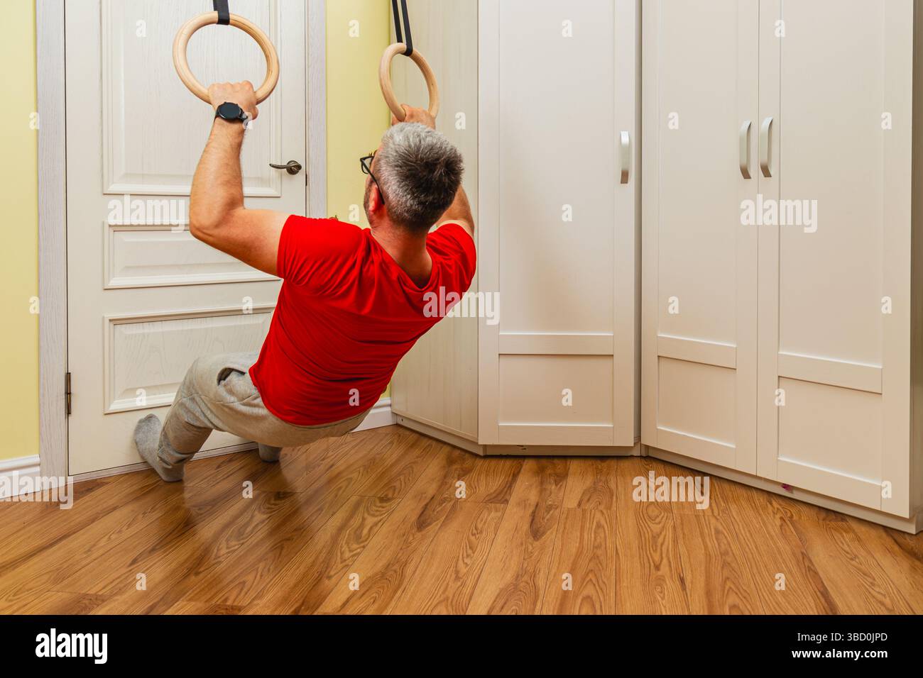 Un uomo esegue una routine di allenamento utilizzando anelli da ginnastica in una stanza ben illuminata. Il pavimento in legno e il design minimalista creano un'atmosfera mirata per gli esterni Foto Stock