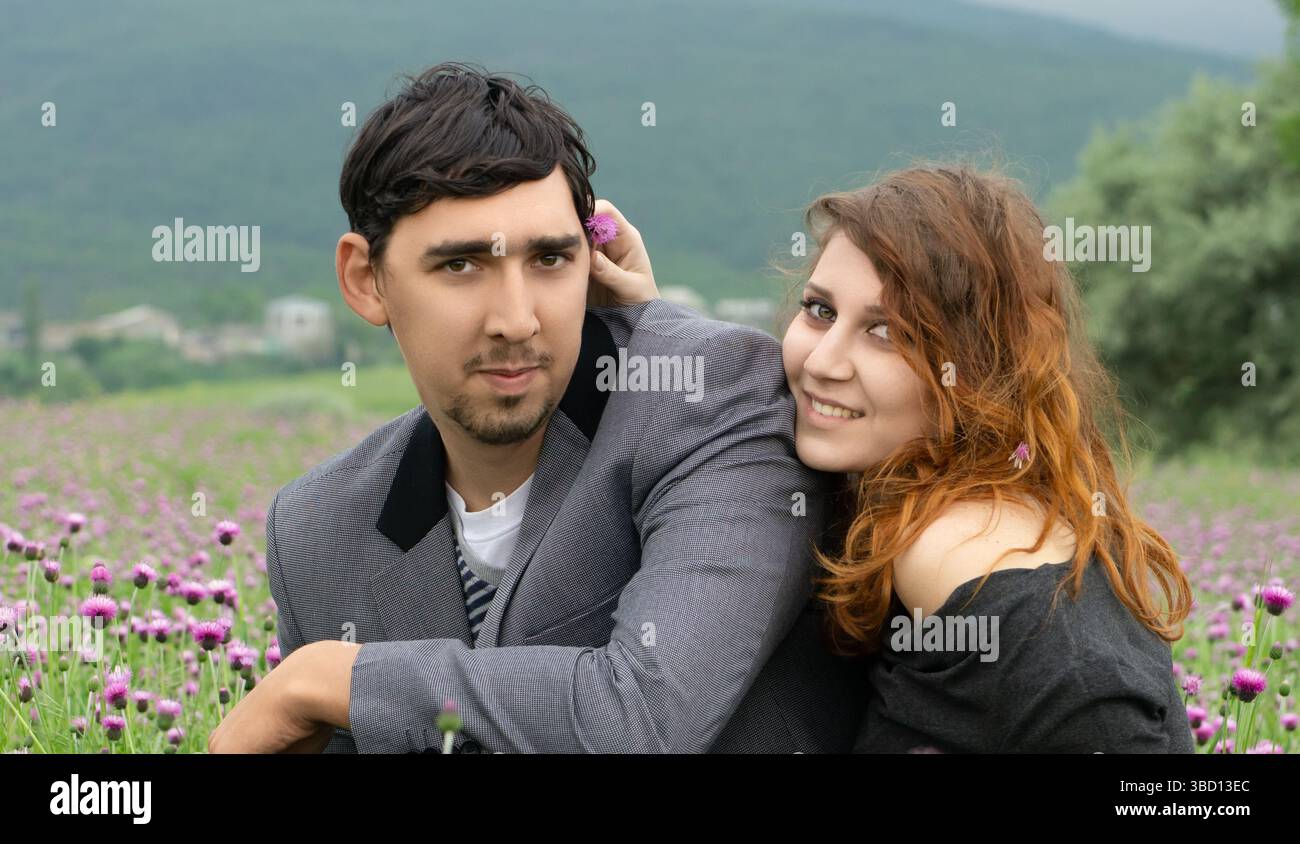 Coppia felice che si gode un momento romantico in un campo fiorito. La donna sorride scherzosamente mentre posiziona un fiore dietro l'orecchio dell'uomo. Foto Stock