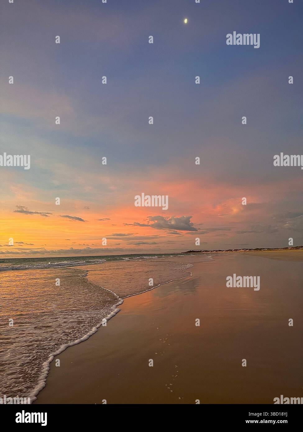 Spiaggia tranquilla al tramonto con cieli pastello, onde morbide e la luna che si innalza: Un tranquillo momento costiero perfetto per viaggi e benessere - Immagine stock catturata con smartphone
