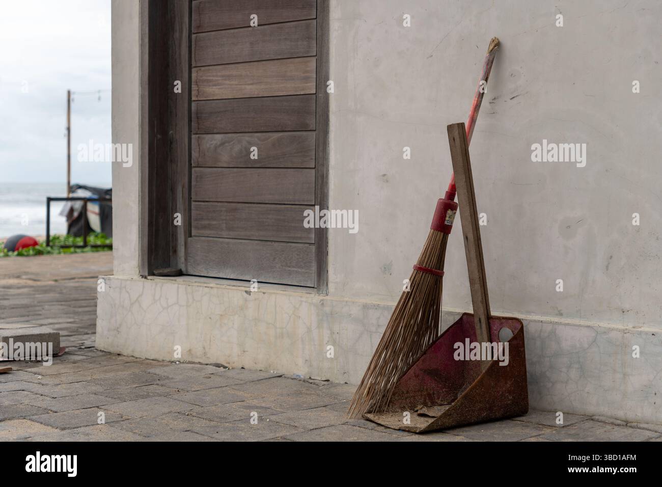 La scopa e una padella sono appoggiate contro un muro. La scopa è rossa e la padella è marrone. La scena è ambientata contro un muro e c'è una spiaggia nella Foto Stock