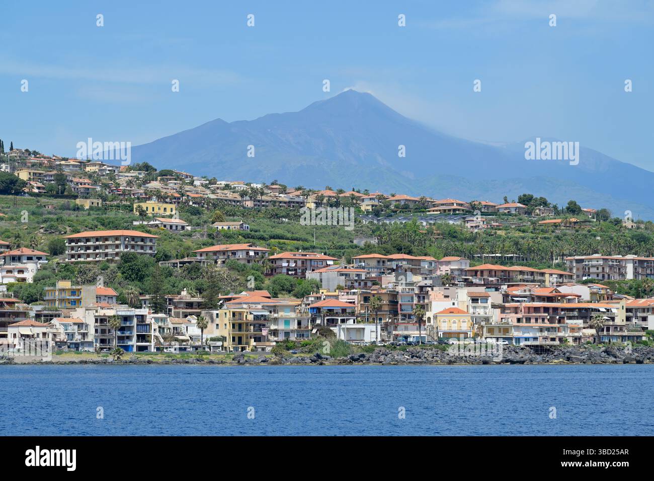 Costa di Aci Castello. Costa dal punto di vista della barca con il vulcano Etna fumante sullo sfondo contro il cielo blu. Sicilia, Italia. Foto Stock