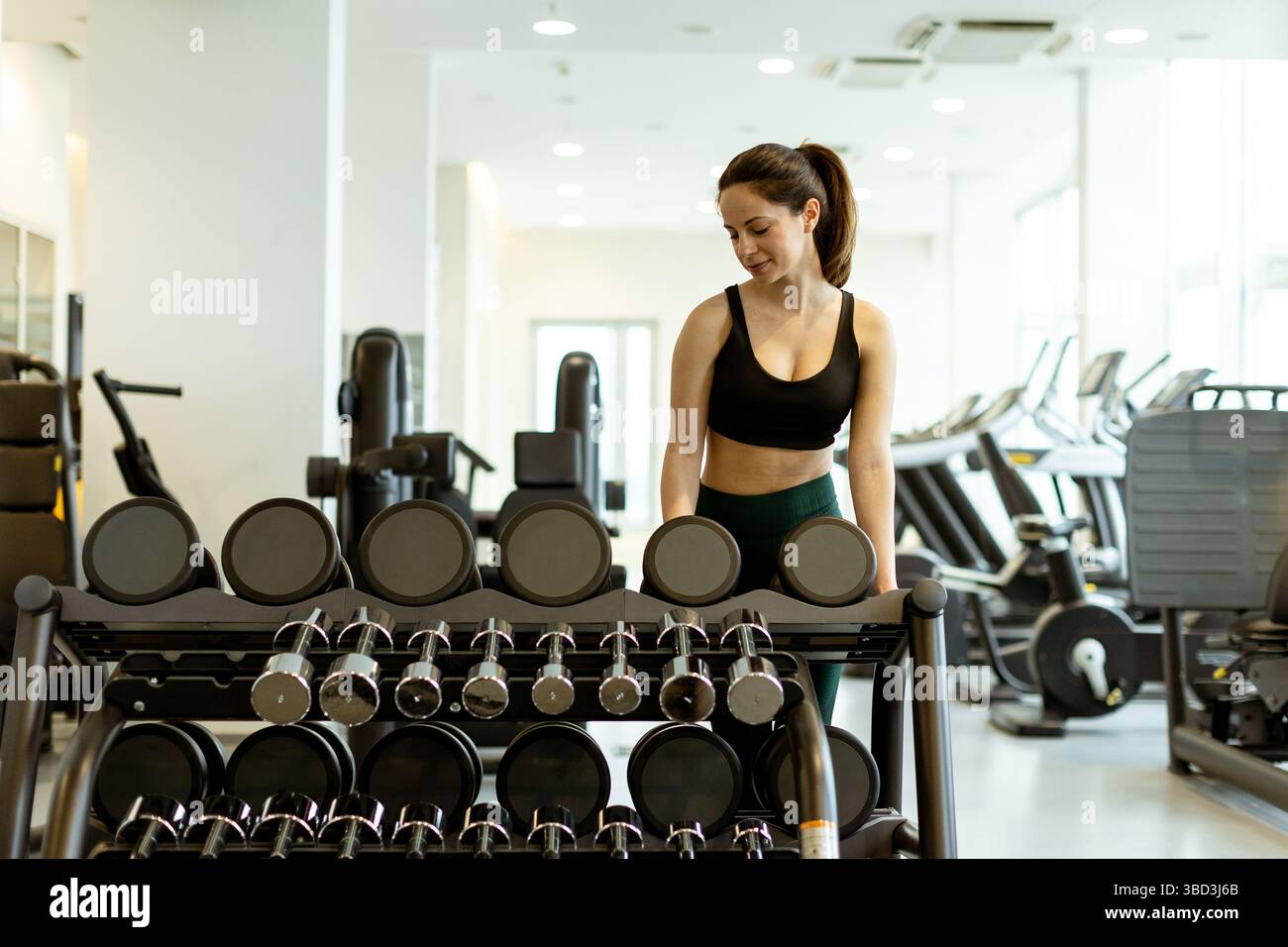 In una palestra contemporanea, una donna in abbigliamento sportivo ispeziona una schiera di manubri, concentrandosi sulla sua routine di allenamento della forza. Foto Stock