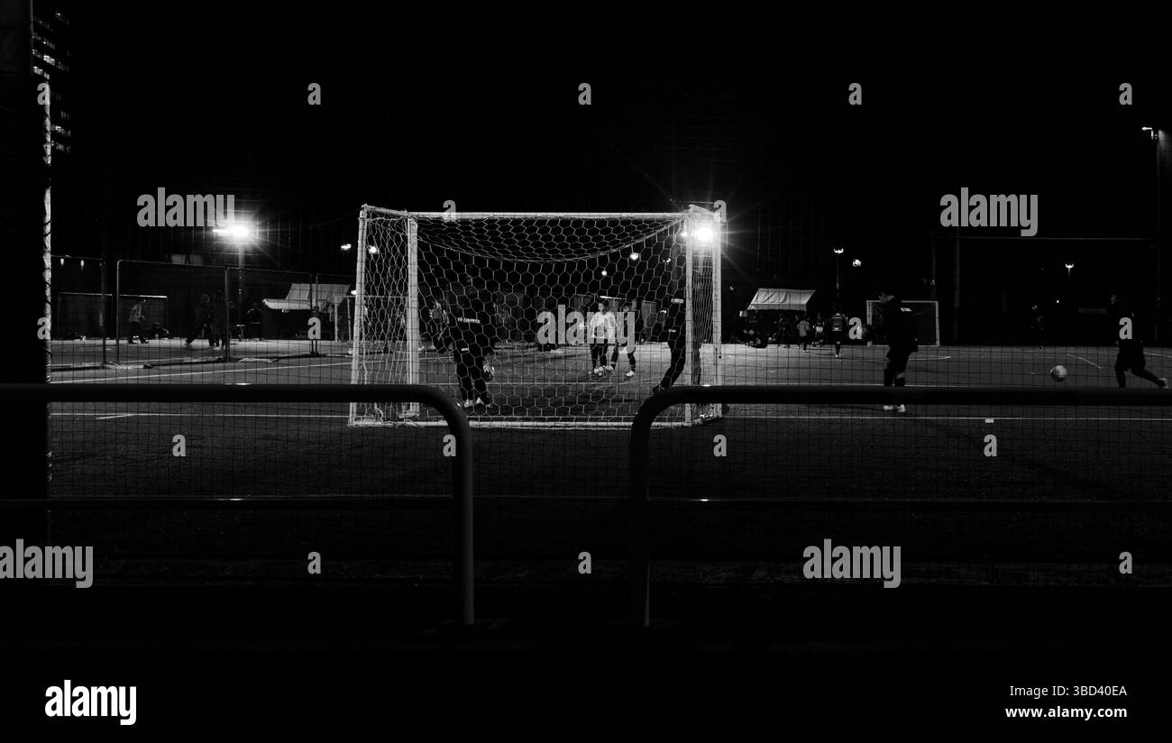 I giovani giocatori si riuniscono per una partita di calcio a tarda notte: Ombre che si estendono sul campo recintato Foto Stock