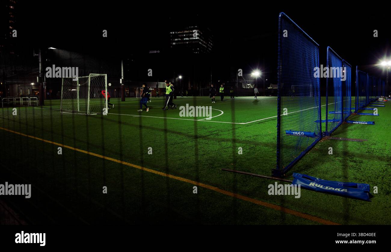 I giocatori inseguono la palla attraverso un campo di erba erbosa, una partita notturna urbana incorniciata da reti e luci della città Foto Stock