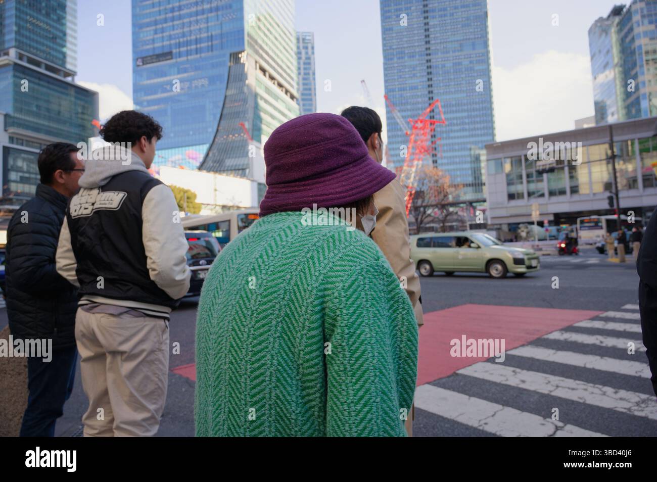 Una donna anziana con un maglione verde e un cappello di prugna aspetta ad un vivace incrocio Foto Stock