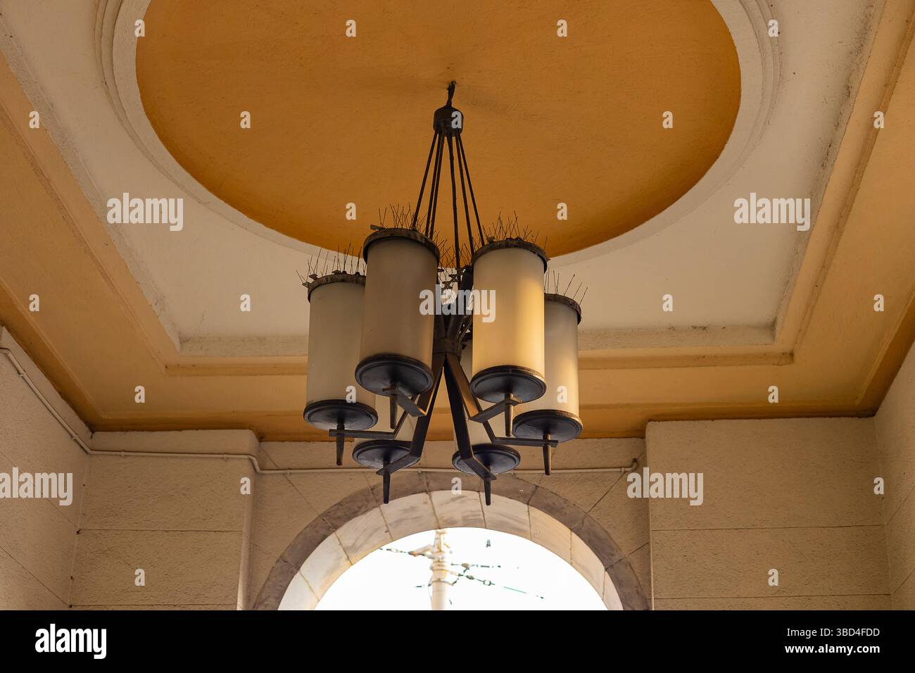 Lampada da soffitto architettonica d'epoca con diverse tonalità cilindriche di vetro e punte deterrenti per uccelli montate in una galleria storica ad arco Foto Stock