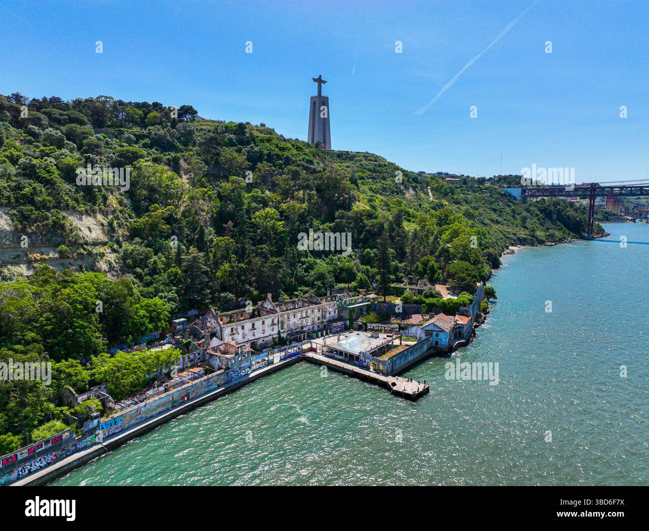 Almada, Portogallo - 12 maggio 2024: Veduta aerea degli edifici abbandonati lungo l'acqua con la statua del Cristo Re in piedi sopra una collina boscosa Foto Stock