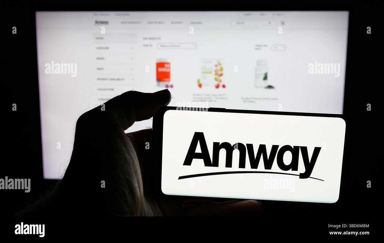 In questa immagine, una persona ha in mano un telefono cellulare con il logo della società di marketing multilivello statunitense Amway Corp. Sullo schermo davanti alla pagina Web. Foto Stock