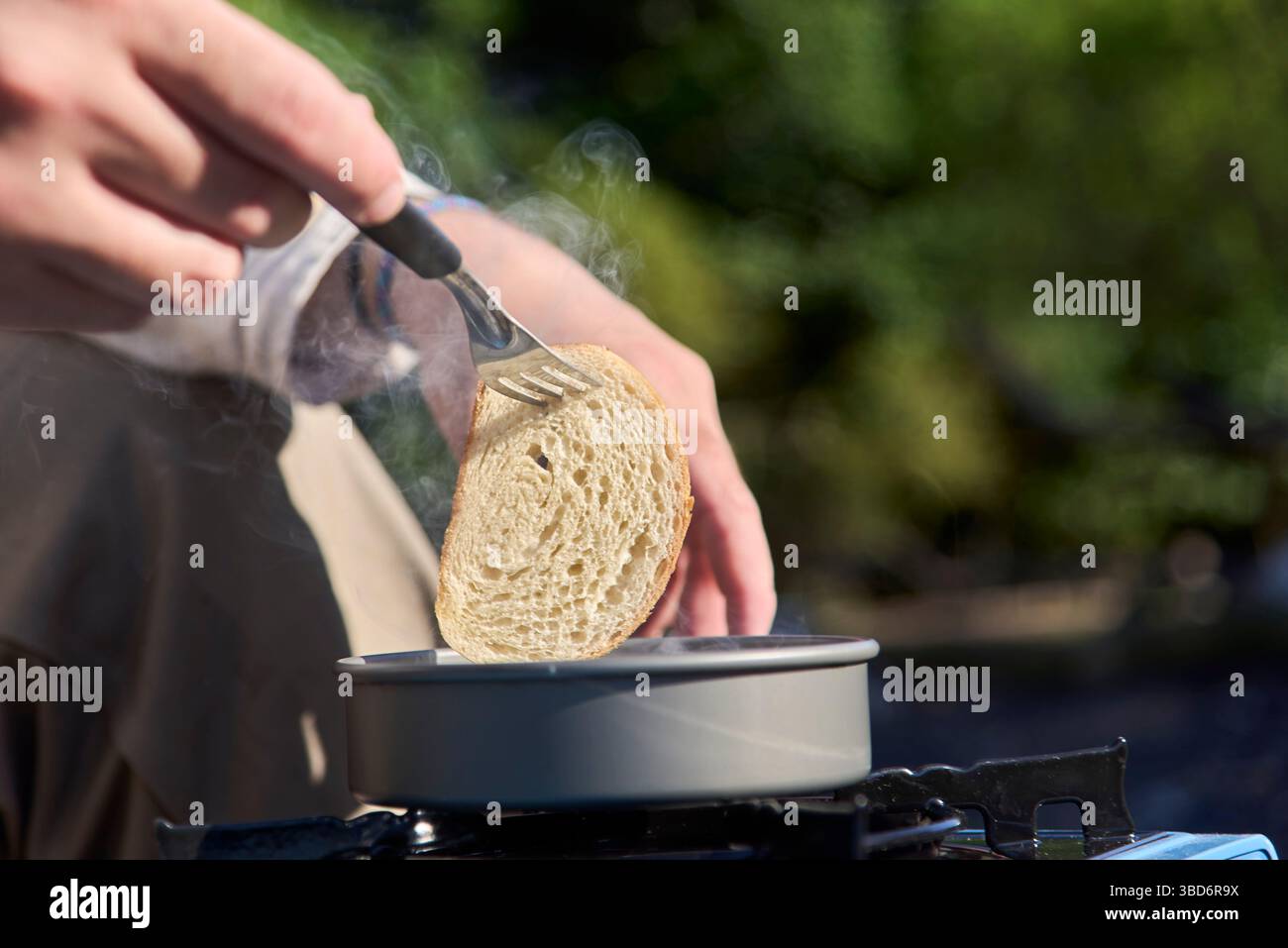 Vista ravvicinata di una fetta di pane tostato sollevata da una persona irriconoscibile con una forchetta da una padella su una stufa portatile, all'aperto. Foto Stock