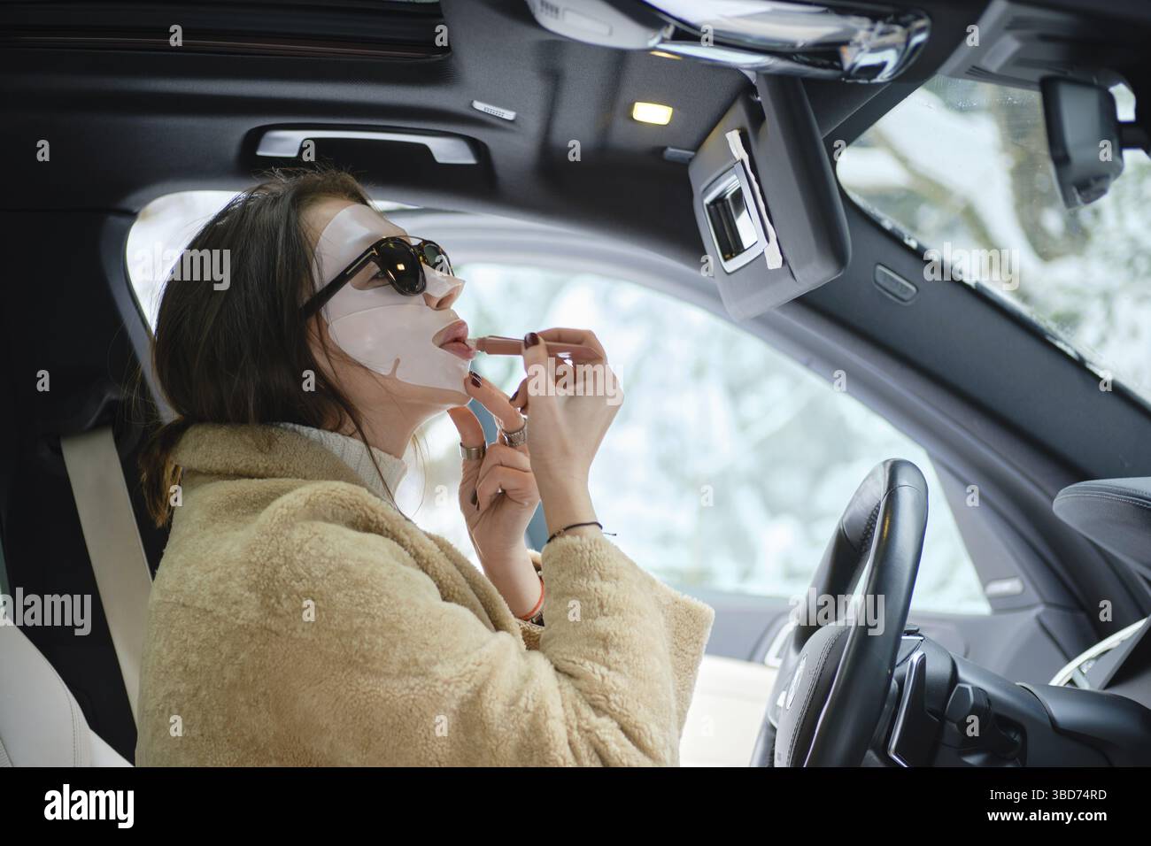 Concetto del ritmo rapido della vita, donna che applica rossetto indossando maschera durante la guida di un'auto Foto Stock