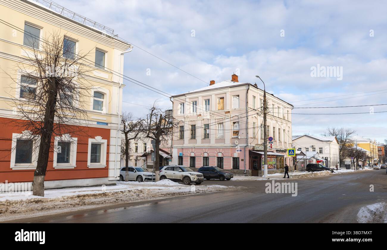 Vladimir, Russia - 5 gennaio 2025: Paesaggio urbano con edifici su una strada innevata durante l'inverno. Veicoli parcheggiati lungo la strada e un pedone Foto Stock