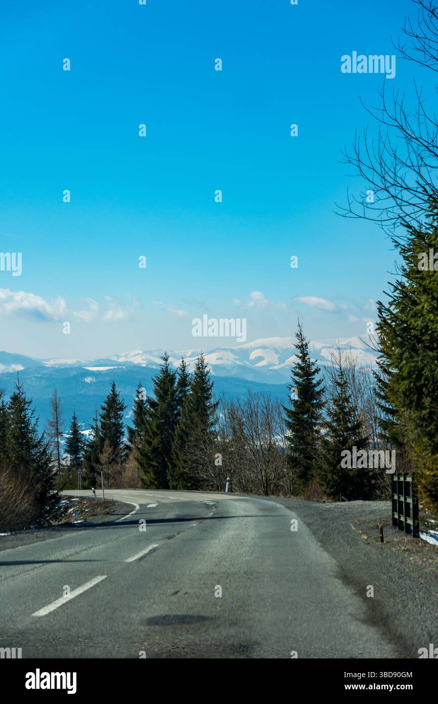 Strada vicino alle montagne. Strada asfaltata con alberi vicino, montagna invernale sullo sfondo. Cielo blu Foto Stock