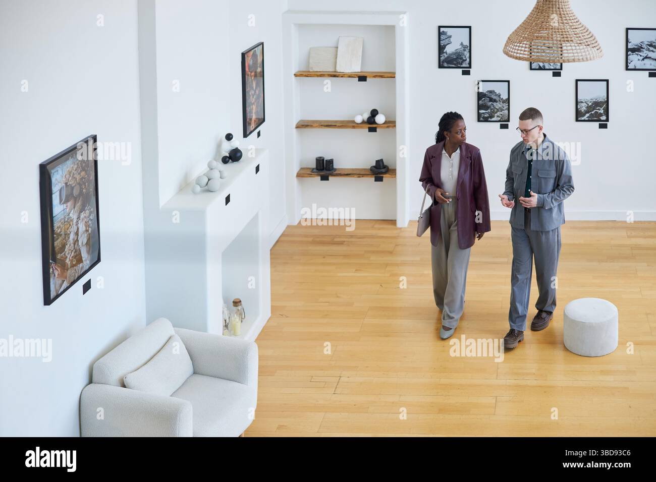 Due persone in piedi in un moderno spazio di galleria, impegnati in una conversazione sul design degli interni. Opere d'arte esposte su pareti e scaffali decorati con va Foto Stock