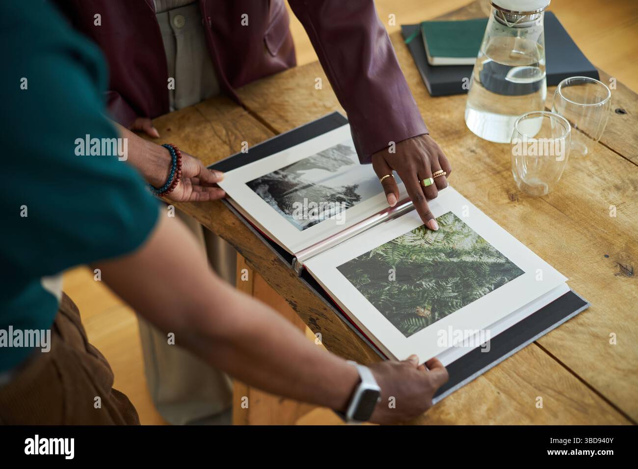 Due persone recensiscono fotografie in un libro aperto su un tavolo rustico in legno, circondato da bottiglia d'acqua e bicchieri. Mani e corpi superiori visibili Foto Stock