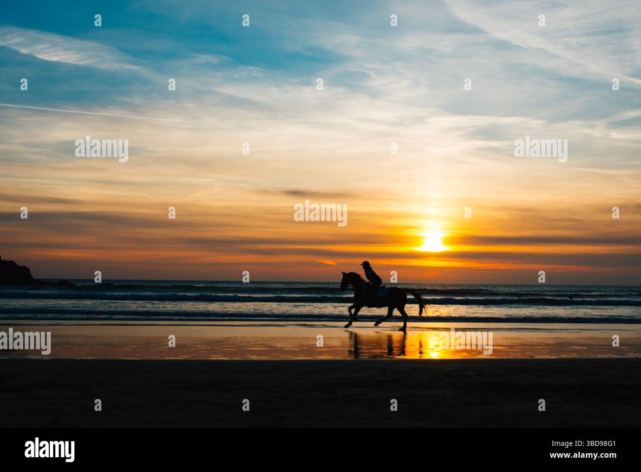 Silhouette di un cavallo e di un cavaliere sulla spiaggia di Perranporth, Cornovaglia, al tramonto, con riflessi dorati sulla sabbia bagnata e le onde che si infrangono dolcemente. Foto Stock
