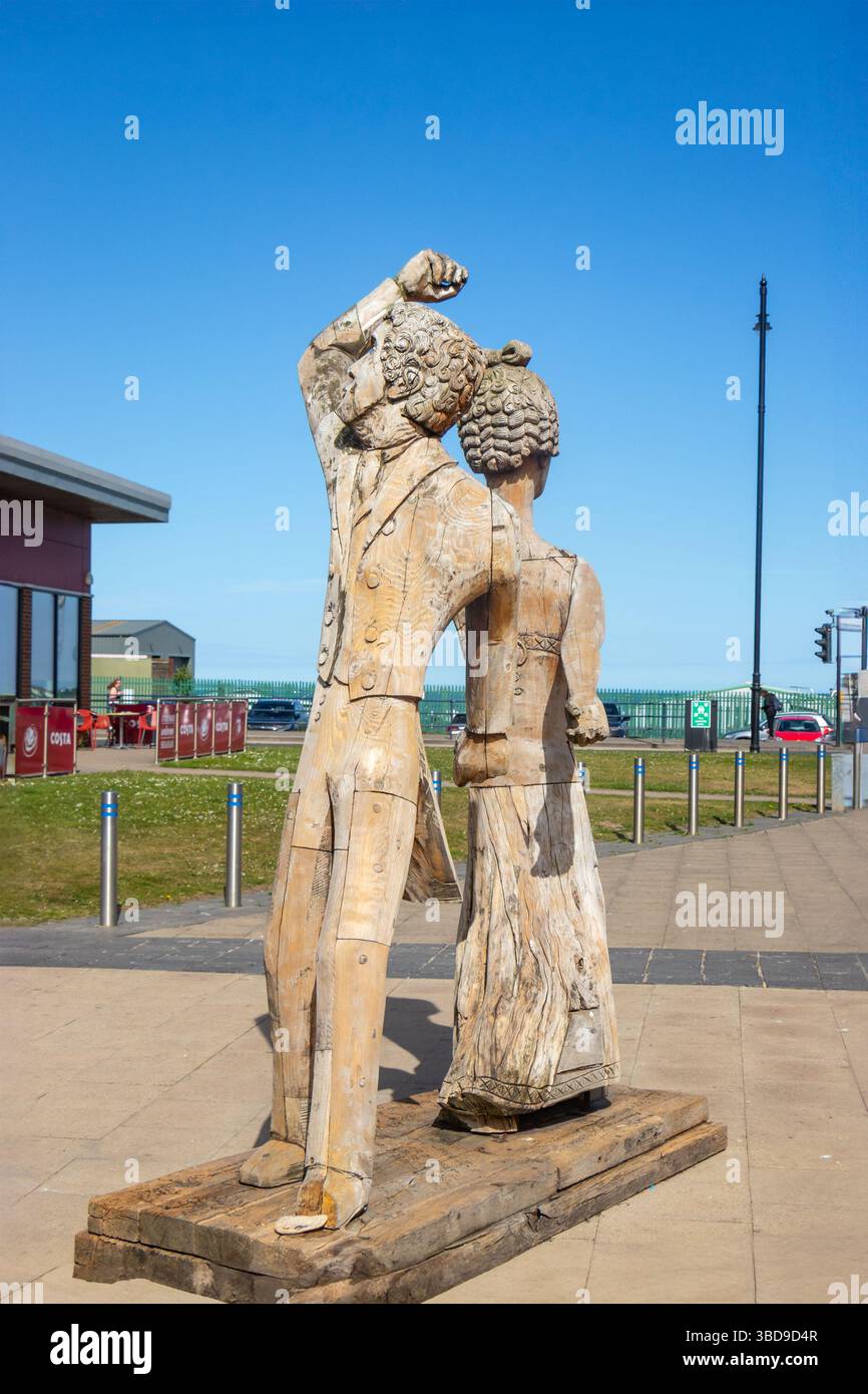 Scultura in legno di una coppia danzante su una passeggiata pavimentata con un cielo azzurro e edifici moderni sullo sfondo Foto Stock
