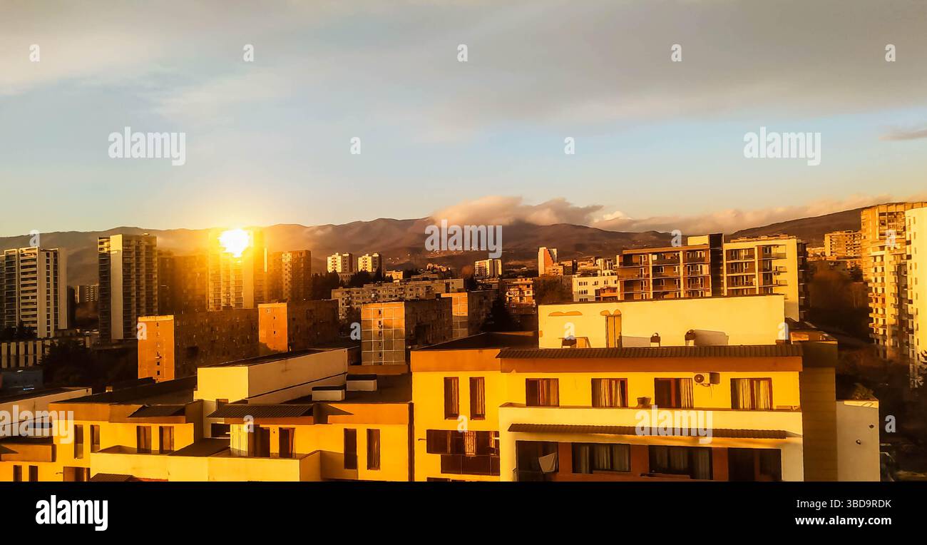 Skyline urbano al tramonto a Tbilisi, Georgia, con luce dorata che diffonde toni caldi su edifici residenziali e montagne sullo sfondo. Pace Foto Stock