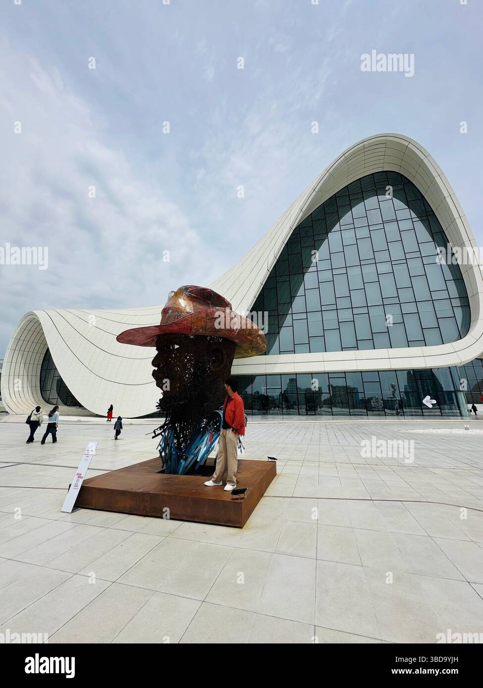 Una moderna scultura di una testa che indossa un cappello da cowboy si erge davanti a un futuristico edificio di vetro a forma d'onda, creando una miscela di arte e cultura Foto Stock