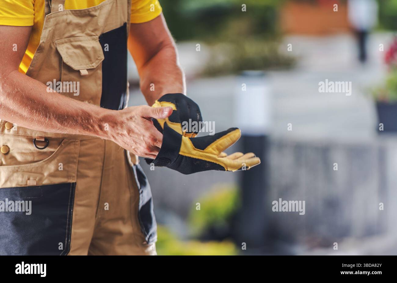 Un operaio edile indossa guanti di sicurezza, preparandosi per un'attività in una vivace area di lavoro all'aperto piena di vegetazione e attrezzi. Il lavoratore è concentrato Foto Stock