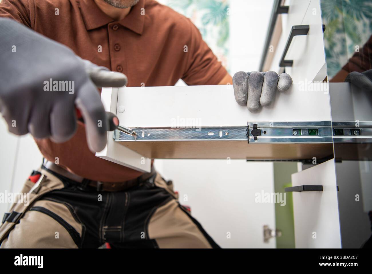 Un lavoratore esperto si concentra sull'installazione di un cassetto dell'armadio, garantendo il corretto allineamento e la funzionalità. La cucina luminosa migliora l'attività Foto Stock