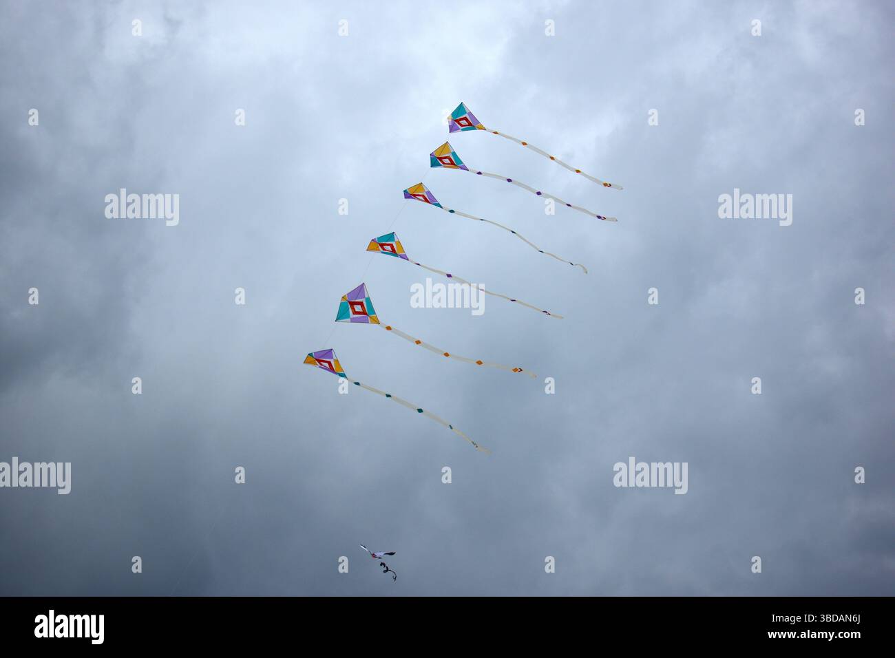Sei coloratissimi aquiloni a forma di diamante con coda posteriore volano in perfetta formazione contro un cielo grigio nuvoloso, creando un contrasto vivido e un senso di movem Foto Stock