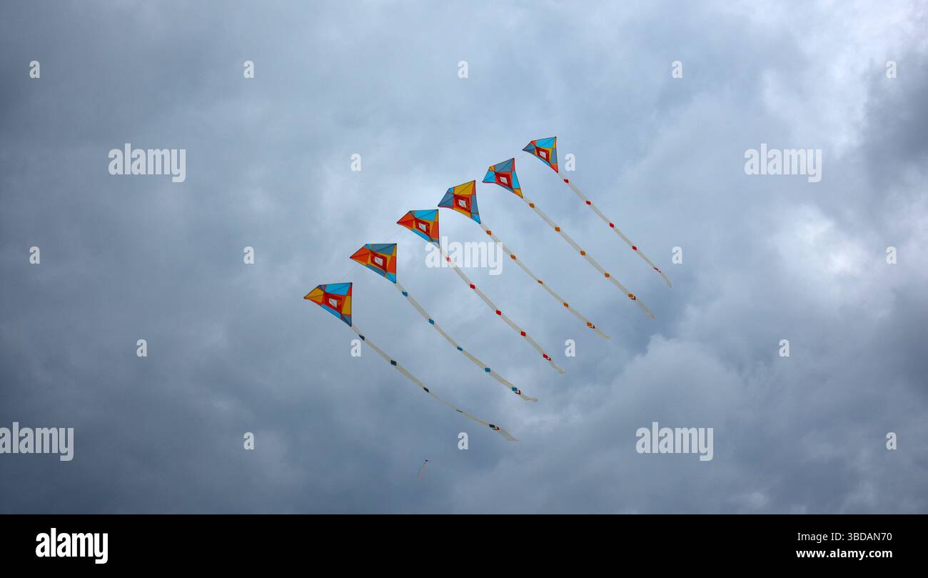 Sei coloratissimi aquiloni a forma di diamante con coda posteriore volano in perfetta formazione contro un cielo grigio nuvoloso, creando un contrasto vivido e un senso di movem Foto Stock