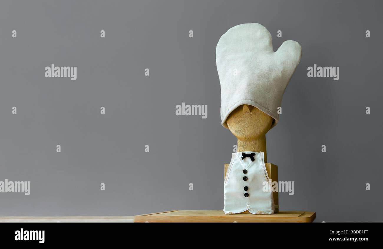 Chef e ristorante. Una miniatura in legno con grembiule di lino e un guanto da cucina in lino al posto di un cappello. Sfondo grigio. Industria alimentare Foto Stock