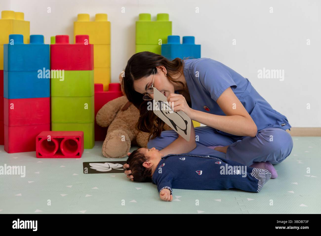 Pediatra che interagisce con un bambino su un tappetino da gioco, circondato da mattoni colorati e giocattoli, promuovendo lo sviluppo della prima infanzia Foto Stock