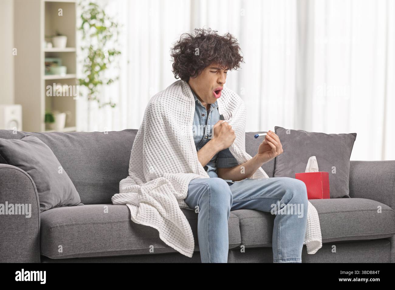 Giovane avvolto in una coperta che si sente male e controlla la temperatura corporea Foto Stock