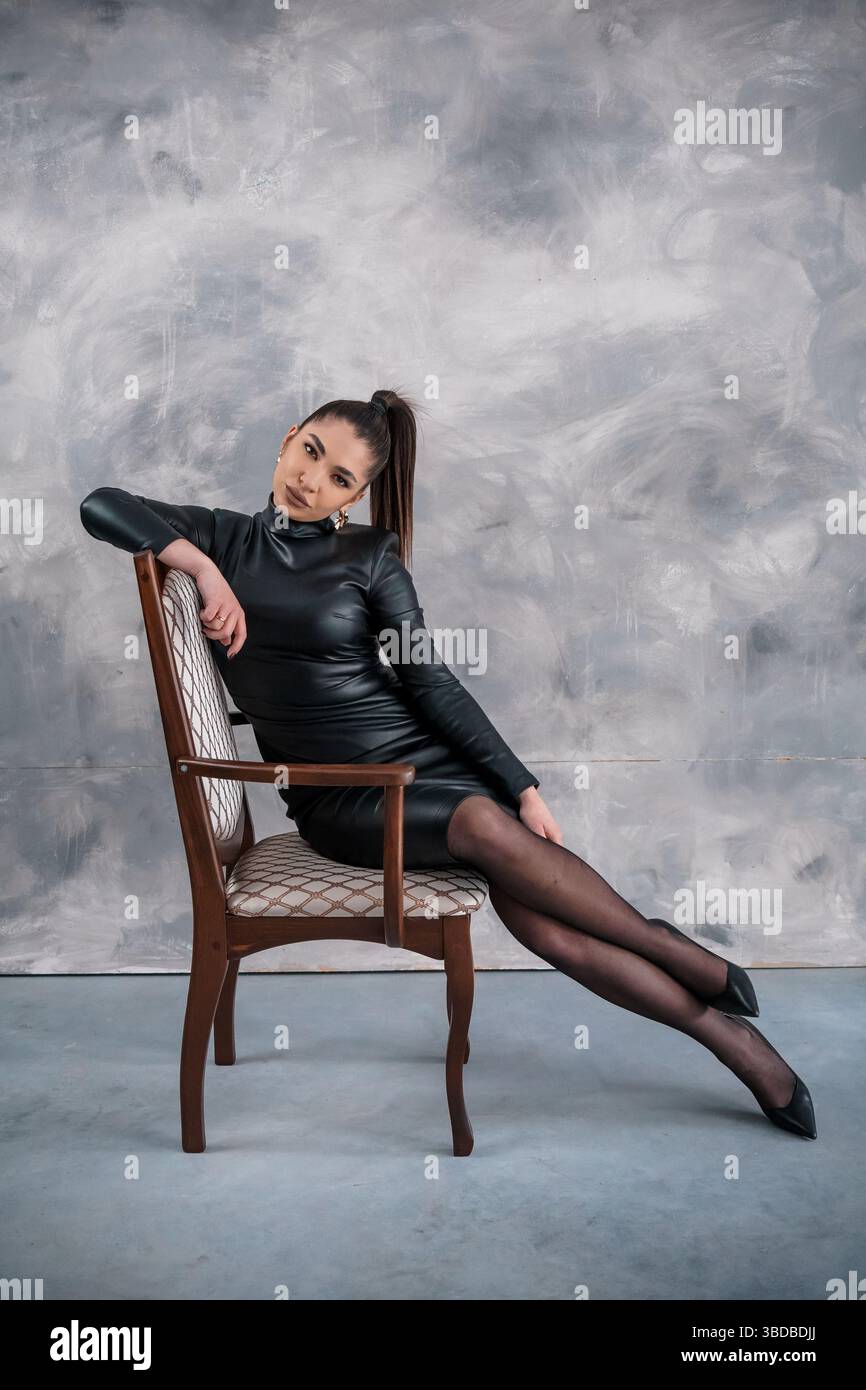 Una giovane donna elegante con i capelli scuri seduta su una sedia di legno, indossa un abito in pelle nera aderente e calze nere, che posa con sicurezza contro una Foto Stock
