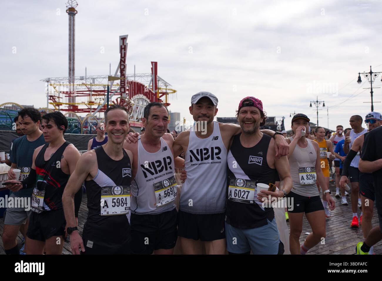 Cinque runner maschili, che indossano un abbigliamento atletico simile e pettorali da corsa, si uniscono dopo la gara su una passerella. E' visibile una grande struttura del parco divertimenti Foto Stock