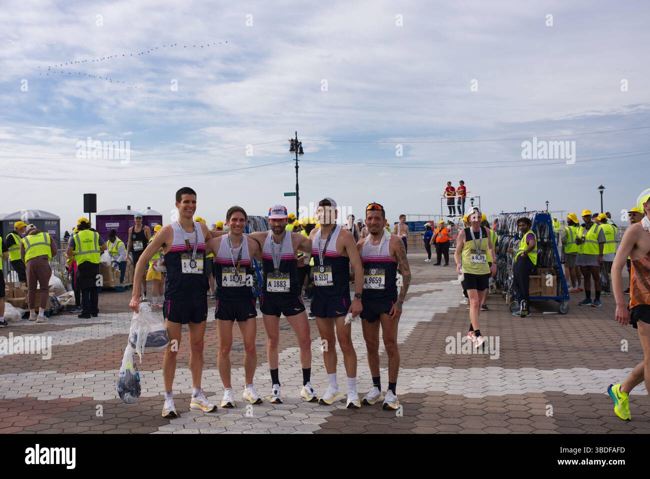 Cinque maratoneti maschili, con bavaglini e medaglie, si uniscono dopo la gara. Sorridono e tengono in mano i loro effetti personali. Spettatori e supp. Gara Foto Stock