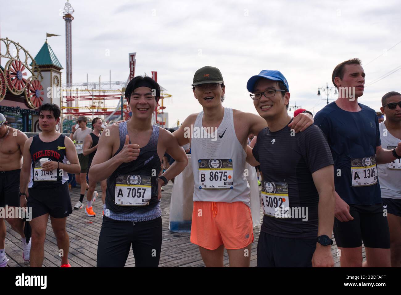 Quattro corridori maschi, che indossano pettorali da gara, posano su una passerella vicino a un parco divertimenti sul mare. Due sorridono e danno i pollici. Gli altri corridori sono VIS Foto Stock